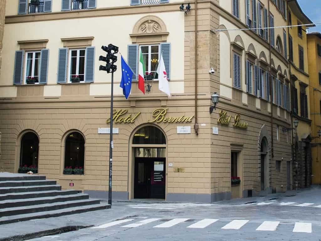 The exterior of The Bernini Palace Hotel on a square right in the cnetre of Florence. There are Italian flags outside the light beige and white stone building. There is also a zebra crossing outside the hotel front door, in Florence.