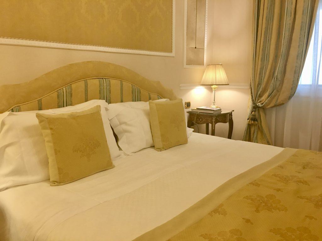 Superior level bedroom with large bed featuring a gold/beige duvet cover, matching cusions and floor to ceiling draping curtains. In the background there is a small white bedside lamp