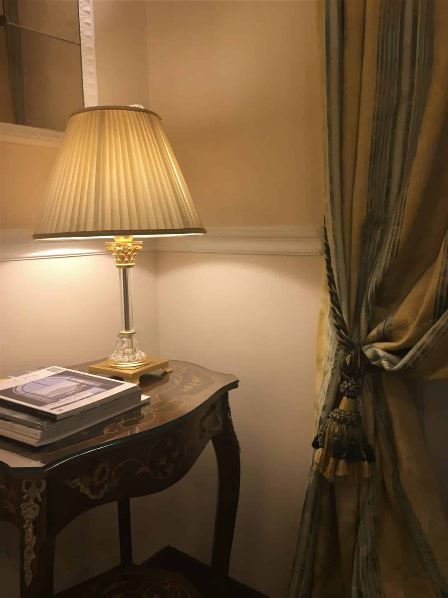 The Opulent Bernini Palace Hotel superior room with a brown bedside table and lamp which is one and bright through the pale green lampshade. There are 2 books on the table and in the corder there is the ceiling to floor long pale green and olive green draped curtains with ties backs