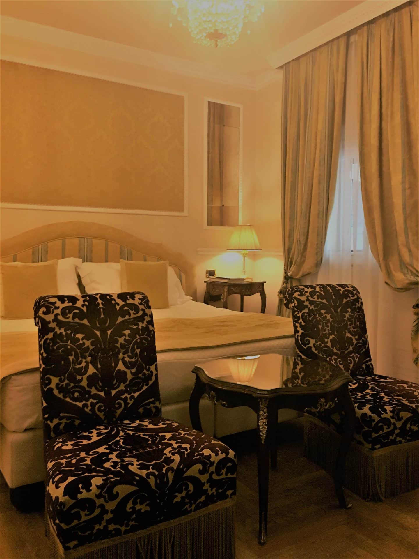 An opulent stay at The Bernini Palace Hotel, Florence. TWo floral chairs with a tabke in between in the superior room. The bed is behind with a stripey olive green adn cream headboard and cream duvet. The long drapey curtains match the headboarc. 