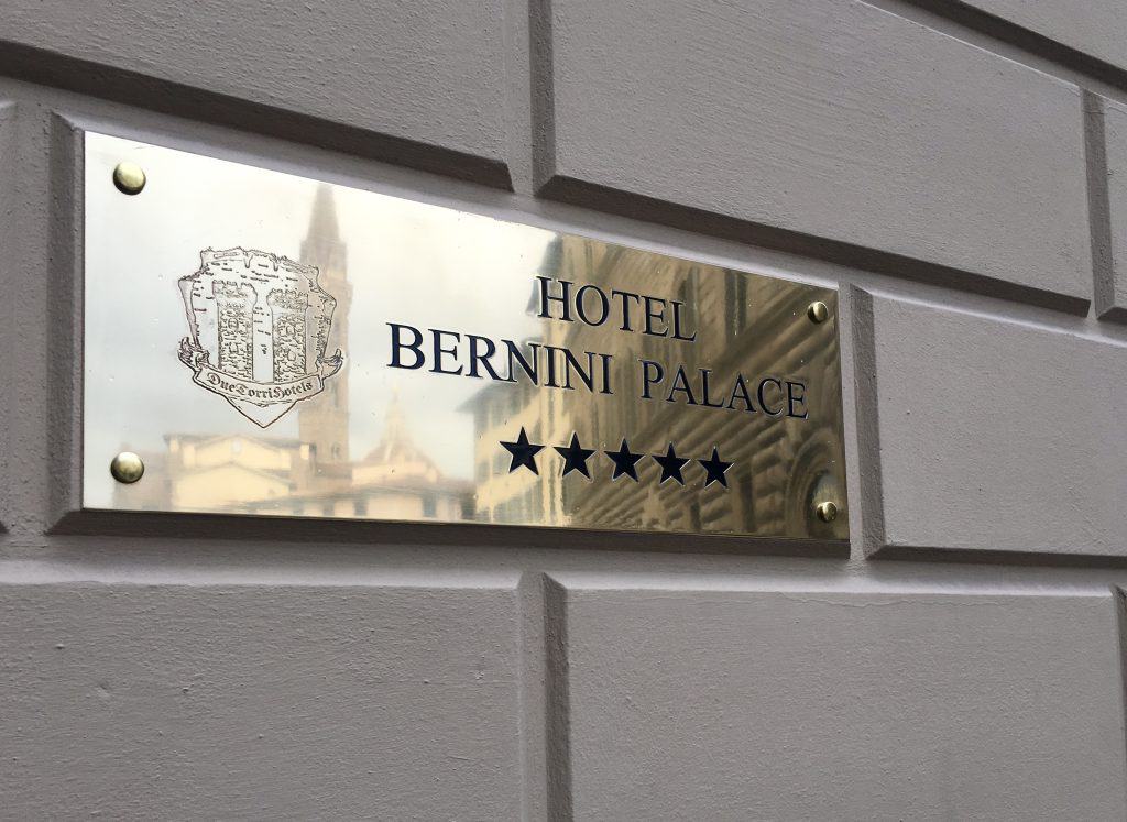A gold plaque utside the Bernini Palace Hoit's name and 5 black stars underneath. the plaque is on top of the beige stone work of the exterior of the hotel.