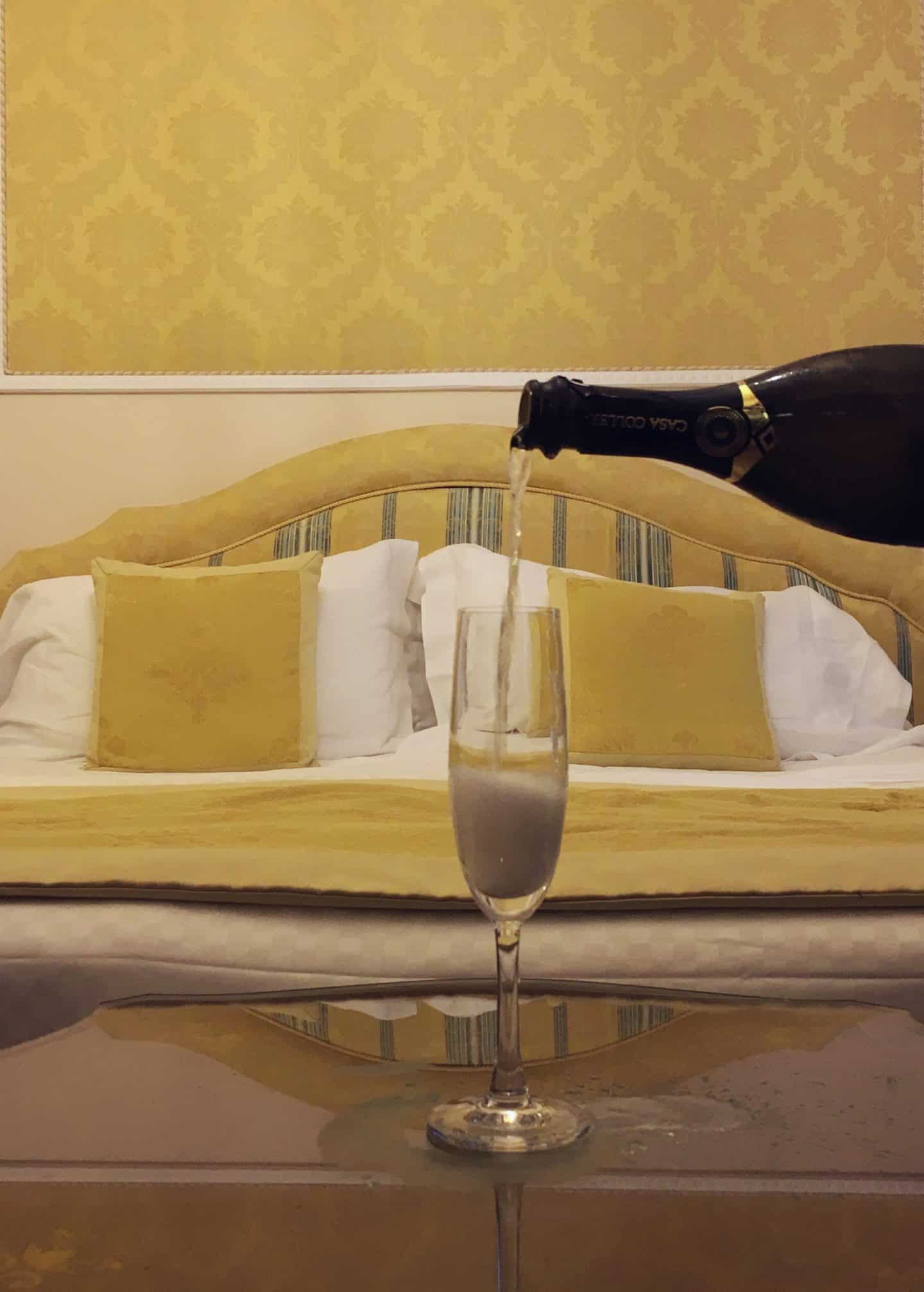 An opulent stay at The Bernini Palace Hotel, Florence. A flue of champagne resting on the dark wood table behind which is a kind size bed. The bed has cream, blue and olive green striped headboard, gold embossed wallpaper and white duvet covers.
