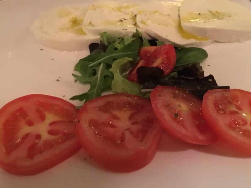 A simple caprese salad with sliced red tomatoes, mozzarella in the back and rocket in the middle. The mozzarella has a drizzling of olive oil. At Il Barretto