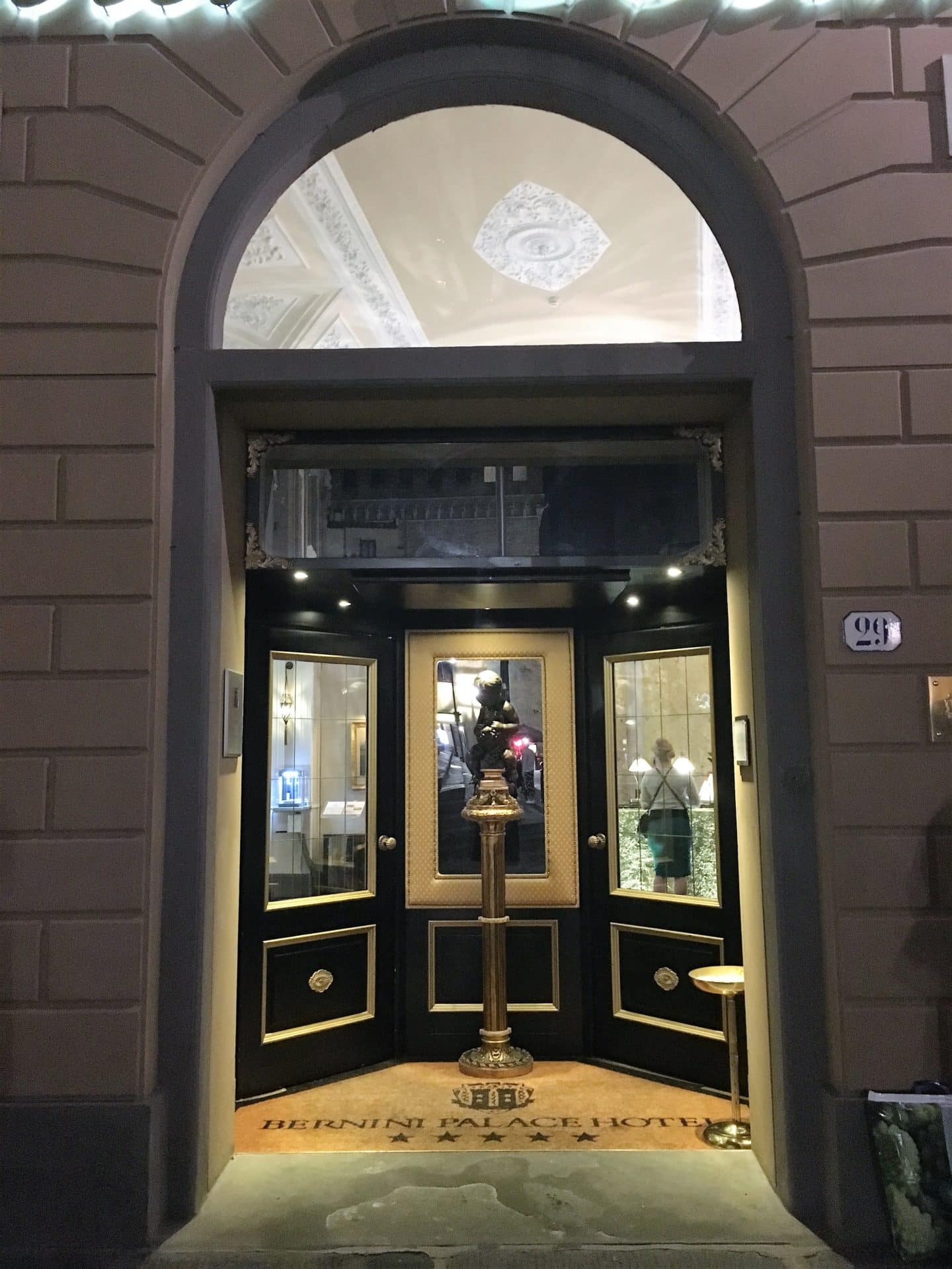 The Opulent Bernini Palace Hotel front door which is dark and split into three walkways. The door is glass and set into the beige stone work of the building. There is a tall flower vase in front of the door, the lights are shininhg through the door