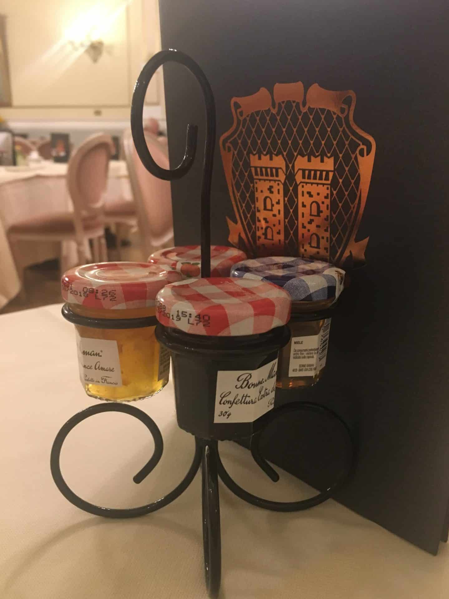 An opulent stay at The Bernini Palace Hotel, Florence. Black wire conserve holder with 4 mini bottle sof jam and marmalade with a dark brown covered Breakfast  menu in the background.