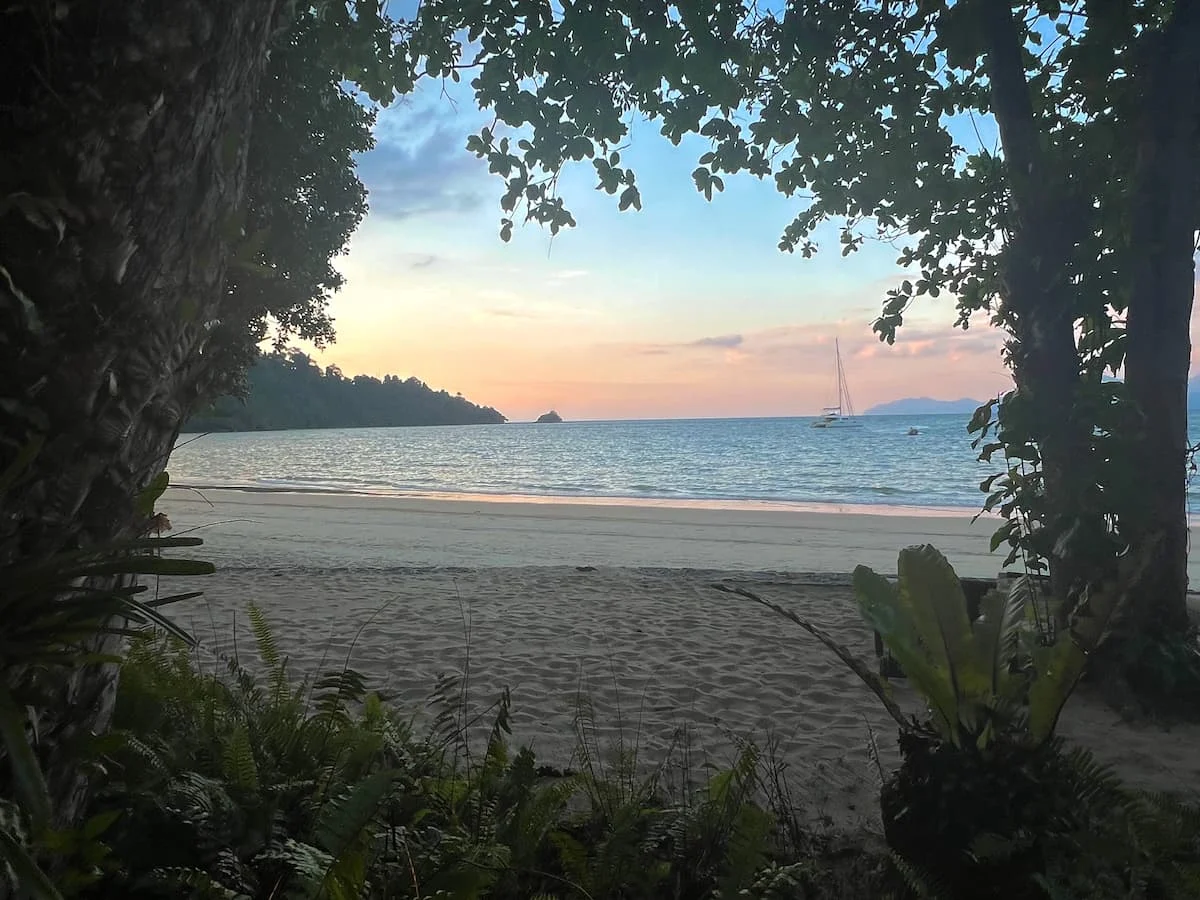 Things to do in Langkawi: Sunset with a Catamaran in the background framed by the green plants of the beach at The Datai, Langkawi